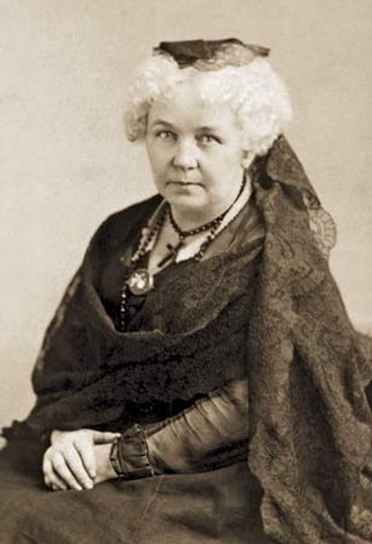 A sepia-toned historical photograph of Elizabeth Cady Stanton, a leading American suffragist and women's rights activist. She is seated, wearing a dark dress, a black lace shawl, and a small dark cap, with white, curly hair and a noticeable necklace with a cross and a medallion.